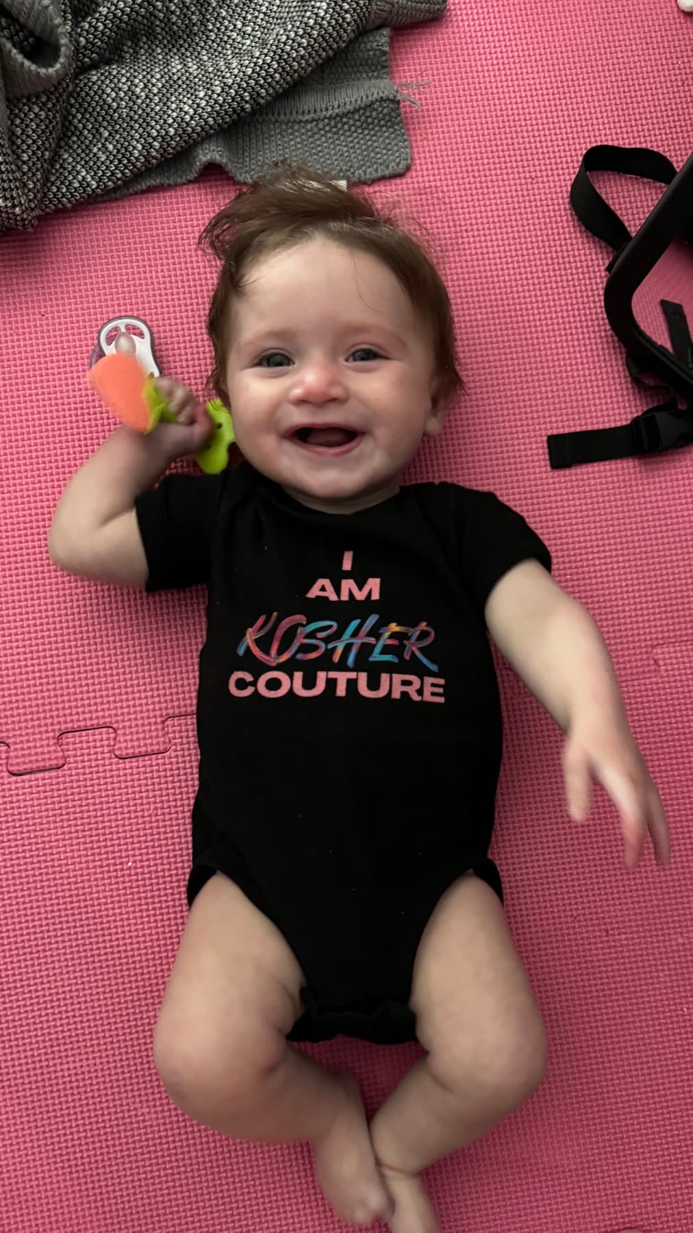 AJ's daughter as a baby, grinning on a play mat in a black I Am Kosher Couture onesie